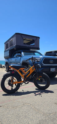 A M7 EILLEO moped-style fat tire electric bike parked in front of a blue Toyota Tacoma with a Tune Outdoor truck camper by the beach.