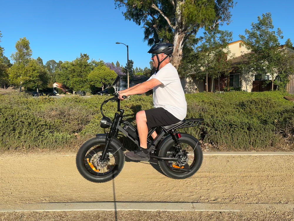 Side view of a man wearing a helmet and sunglasses riding a black EILLEO M6 moped-style fat tire electric bike on a dirt path in a sunny park.