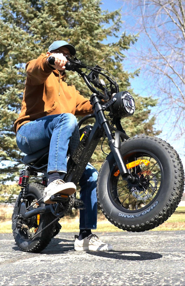 Low-angle action photo of a man in a brown hoodie performing a wheelie on a Eilleo M7 fat tire electric bike